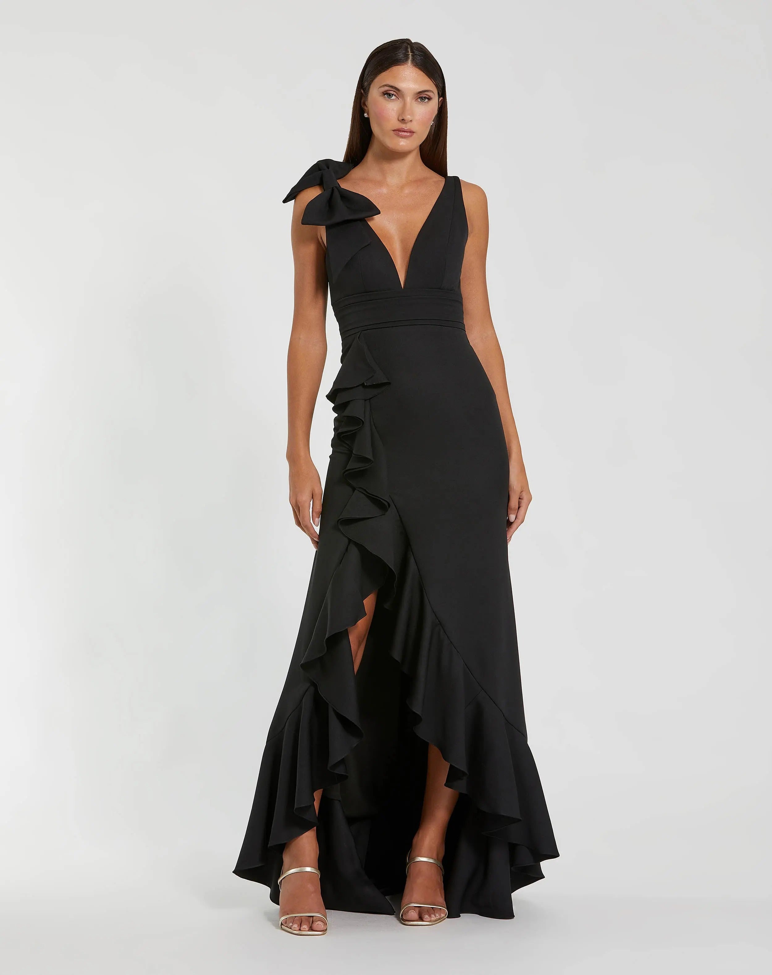 A woman stands against a plain light background, dressed in the Mac Duggal 49965 long formal dress—a sleeveless black evening gown featuring a deep V-neckline, ruffle accents, a side shoulder bow, and a striking high-low hem.